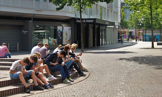 A group of teens sitting on bench and playing on their devices. 